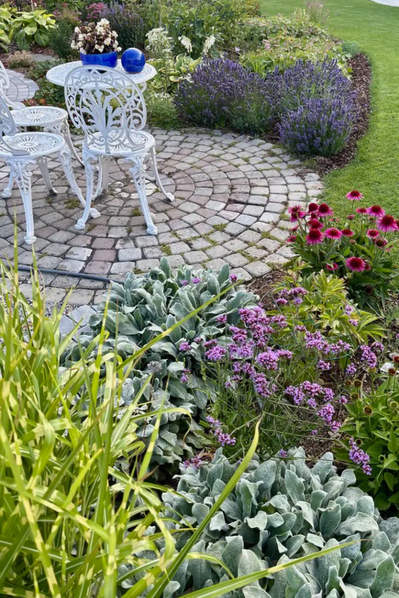 Kleine Sitzgelegenheit in einem mediterranem Garten. 3 weiße Stühle im filigranen Metalldesign, ein weißer Tisch. Alle Elemente stehen auf einem rund angelegtem Pflastersteinboden. Runderherum sieht man verschiedene Pflanzen und Kräuter.