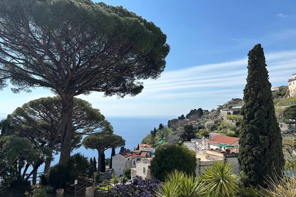 Aufnahme einer kleiner Küstenstadt am Meer an der italienischen Amalfi Küste. Im Vordergrund sieht man Zypresen und Pinien, im Hintergrund Häuser und das Meer.