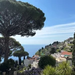 Aufnahme einer kleiner Küstenstadt am Meer an der italienischen Amalfi Küste. Im Vordergrund sieht man Zypresen und Pinien, im Hintergrund Häuser und das Meer.