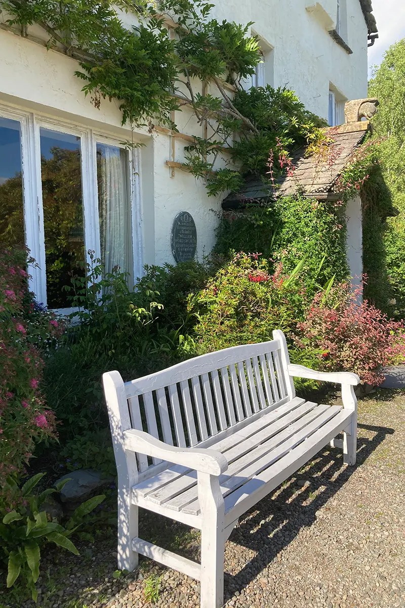 Ausschnitt aus einem Garten im englischen Cottage Garten Stil, man sieht eine schöne weiße Holzbank, die hinter einem Haus steht. Zwischen Haus und Bank sind viele grüne Pflanzen und Büsche zu sehen
