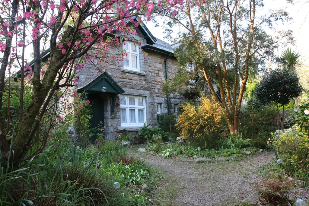Hübsches englisches Cottage im typischen Baustil mit Ziegeln und einem romantischen Cottage Garten mit Pflanzen und Bäumen im Vordergrund.