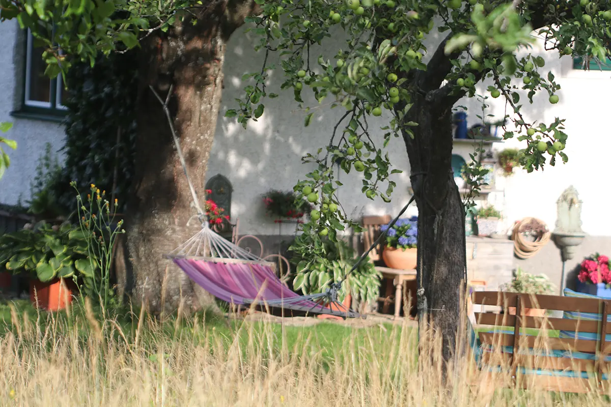 Gemütlicher Garten im Landhausstil. Im Vordergrund sieht man eine Hängematte, die zwischen zwei äumen aufgehängt ist.